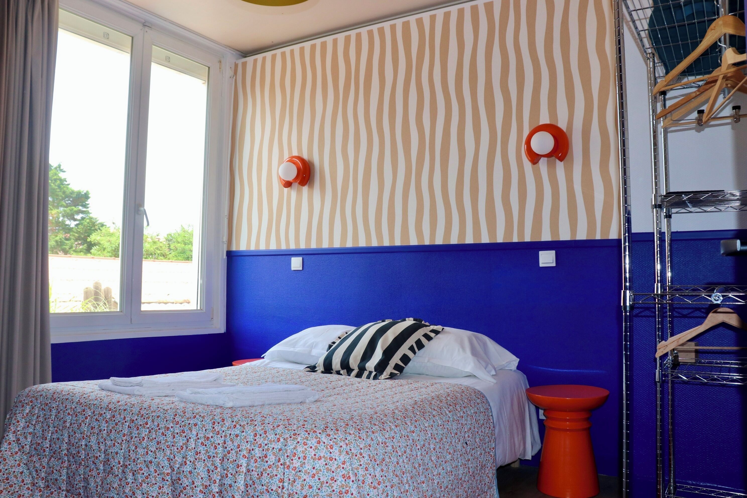 Ein Schlafzimmer mit Bett, Fenster, blauer Wand und rotem Hocker.