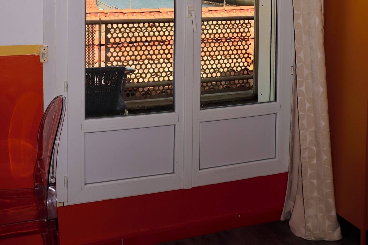 A room with a large window overlooking a beach and a building. There is a chair and a TV mounted on the wall.