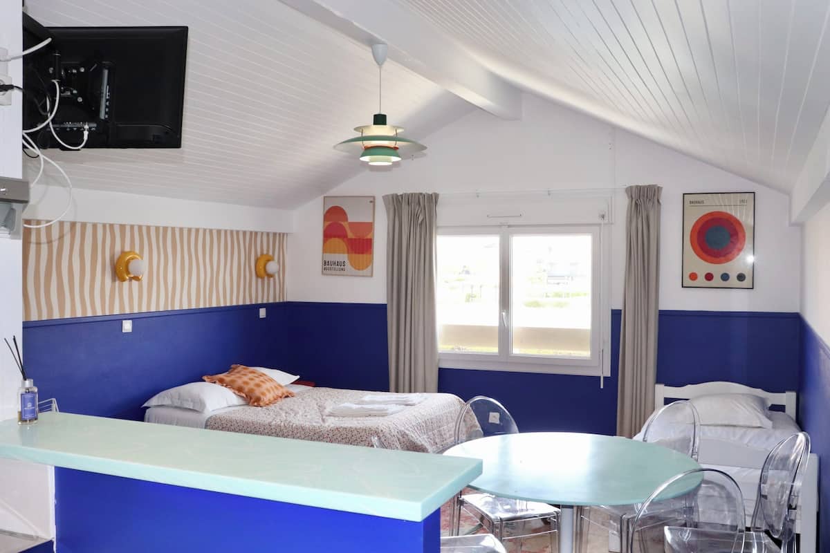 A room with two beds, a small table, and a chair. There is a window with curtains, a mounted television, and a poster on the wall.