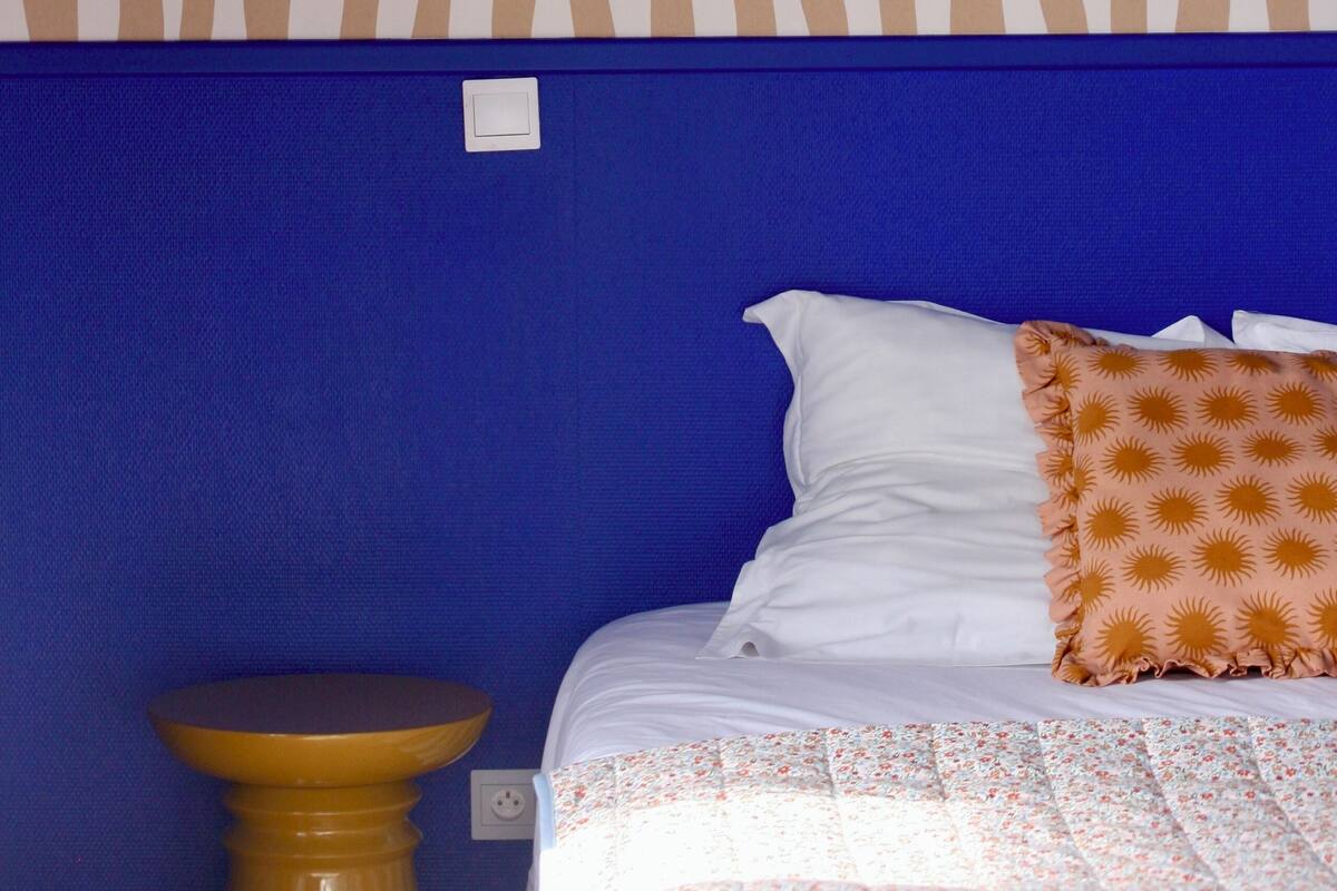 A bedroom with a patterned wallpaper, a blue headboard, a bed with white and patterned pillows, and a small round table.