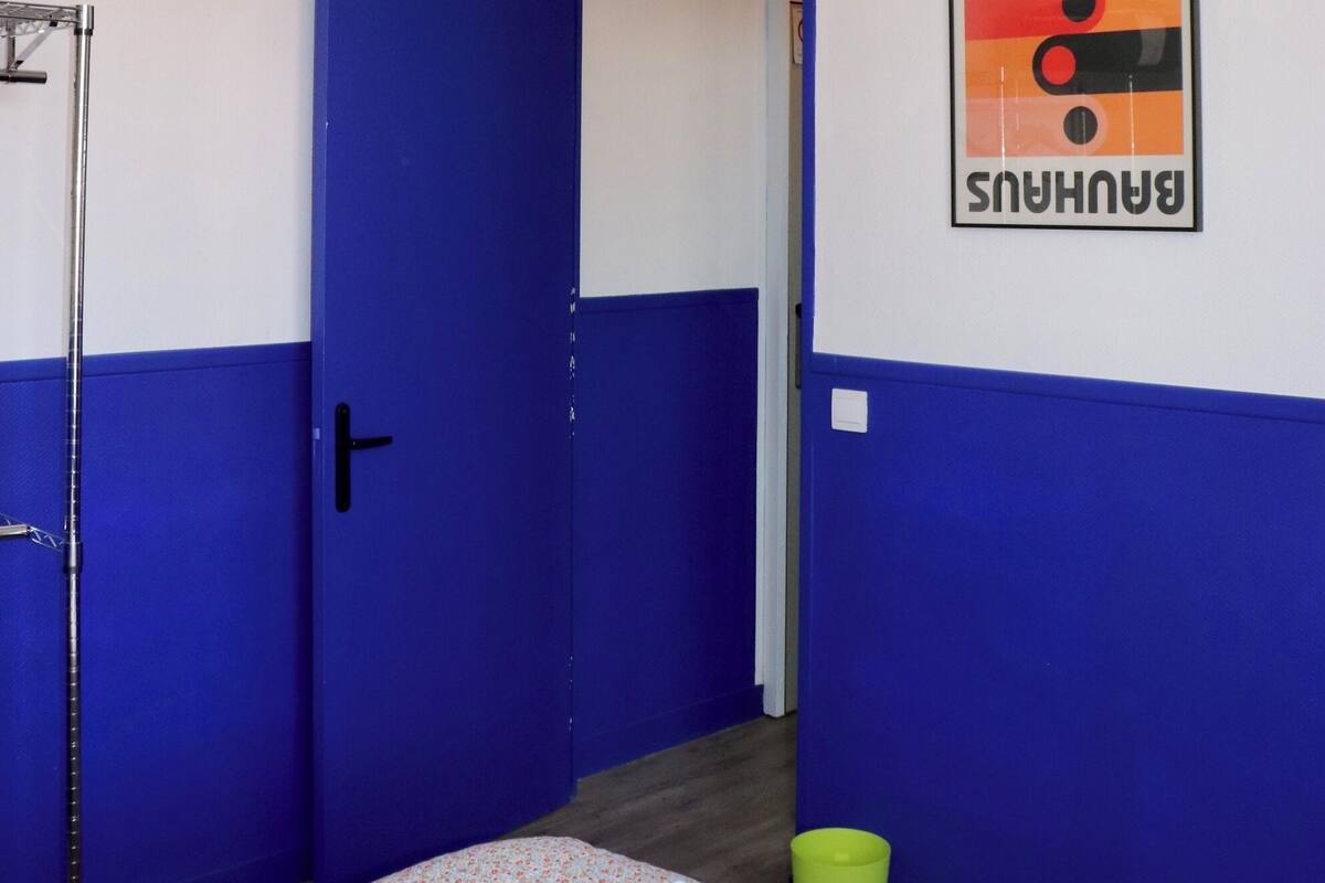 A room with a bed, a yellow hanging light, a blue door, and a Bauhaus poster on the wall.