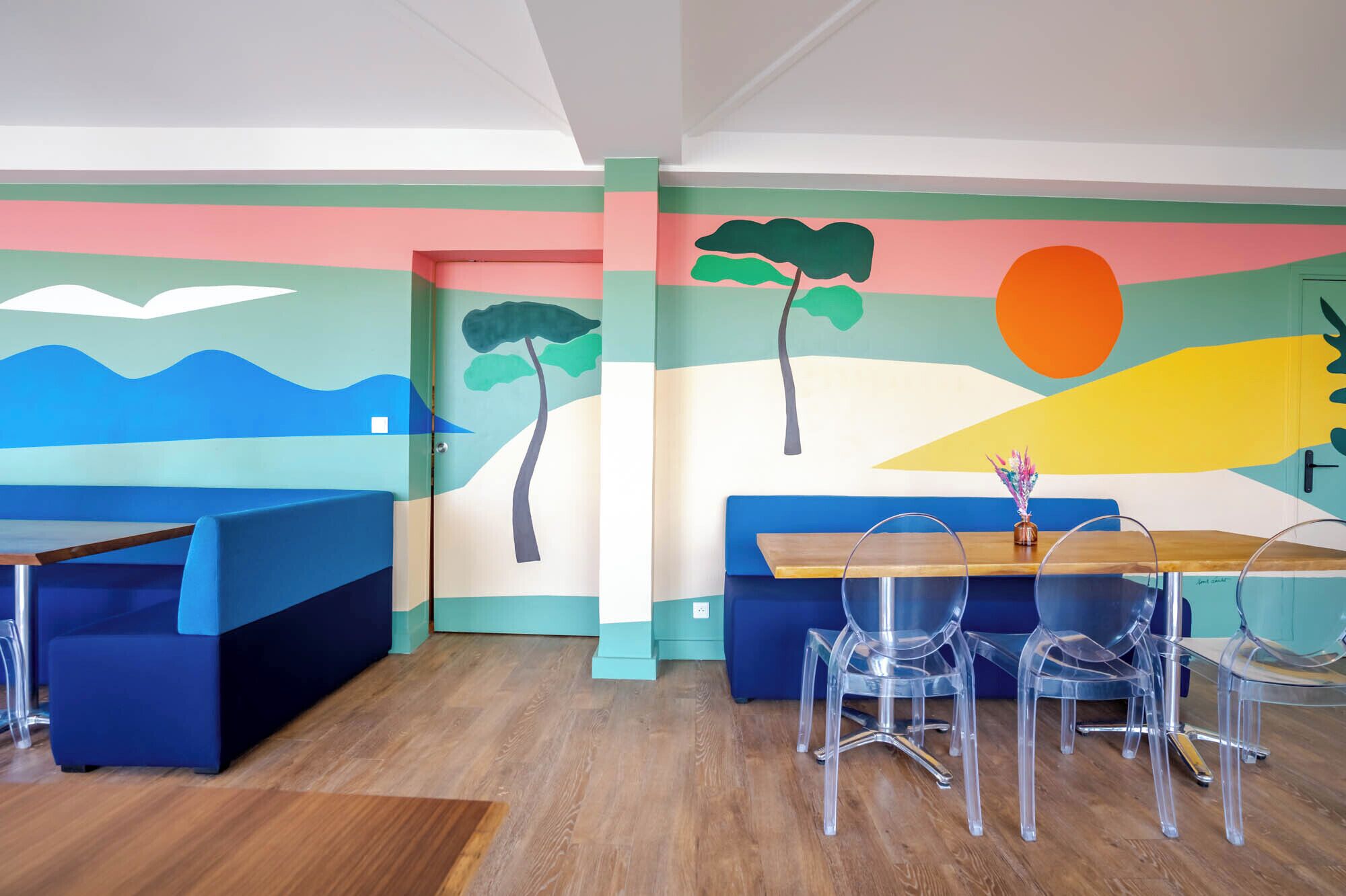 Un salón infantil lleno de color, con asientos azules y verdes, una mesa de madera y un mural de un paisaje con árboles y sol.