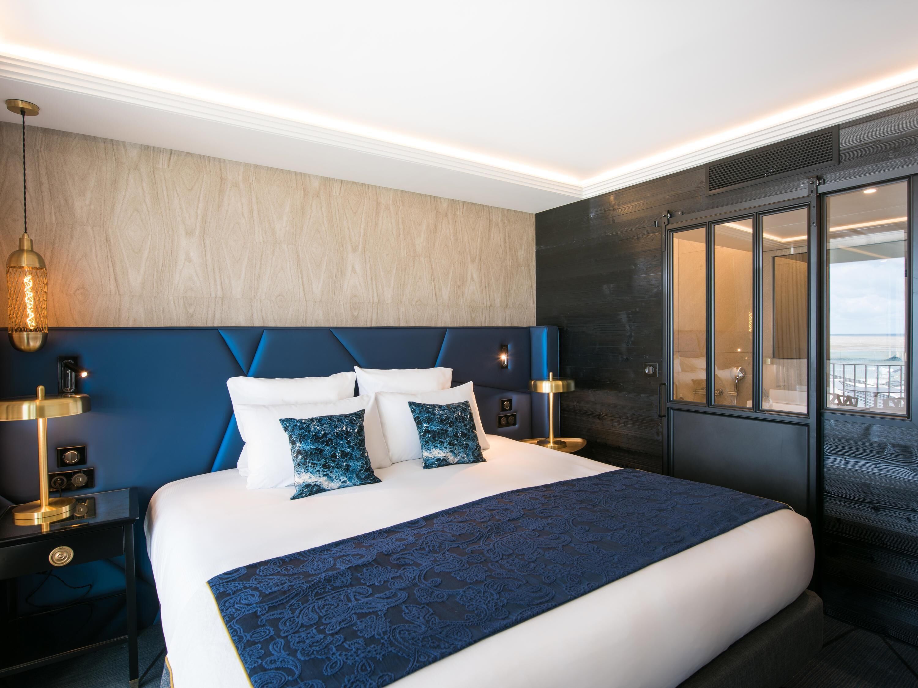 Habitación de hotel moderna con una cama grande, vista al océano y un paisaje urbano visible a través de la ventana.