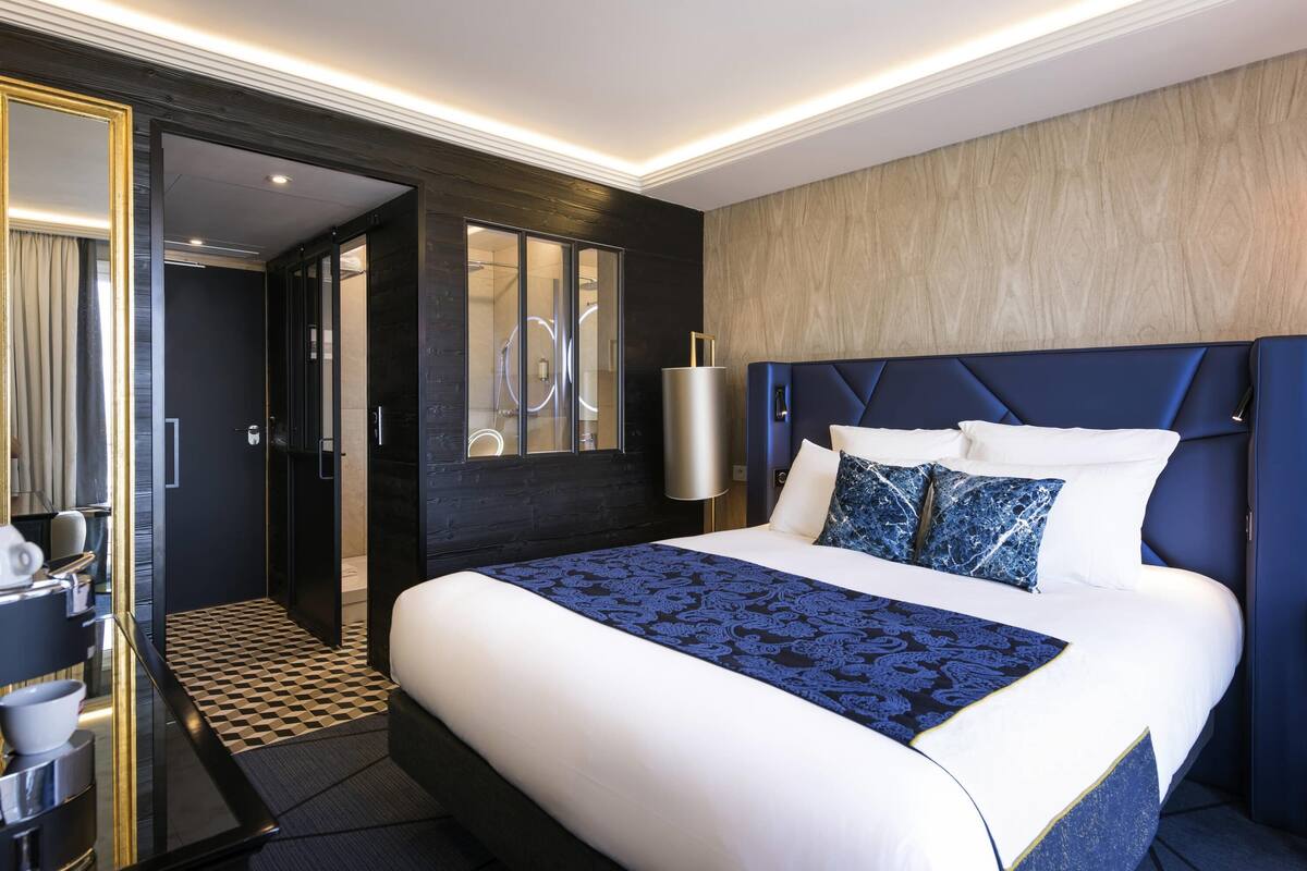 A modern hotel room with a large bed, a dark wooden headboard, a bedside table, and a mirror.