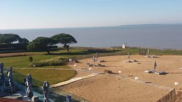 A beach with sun loungers and umbrellas, a grassy area with trees, and a view of the sea.