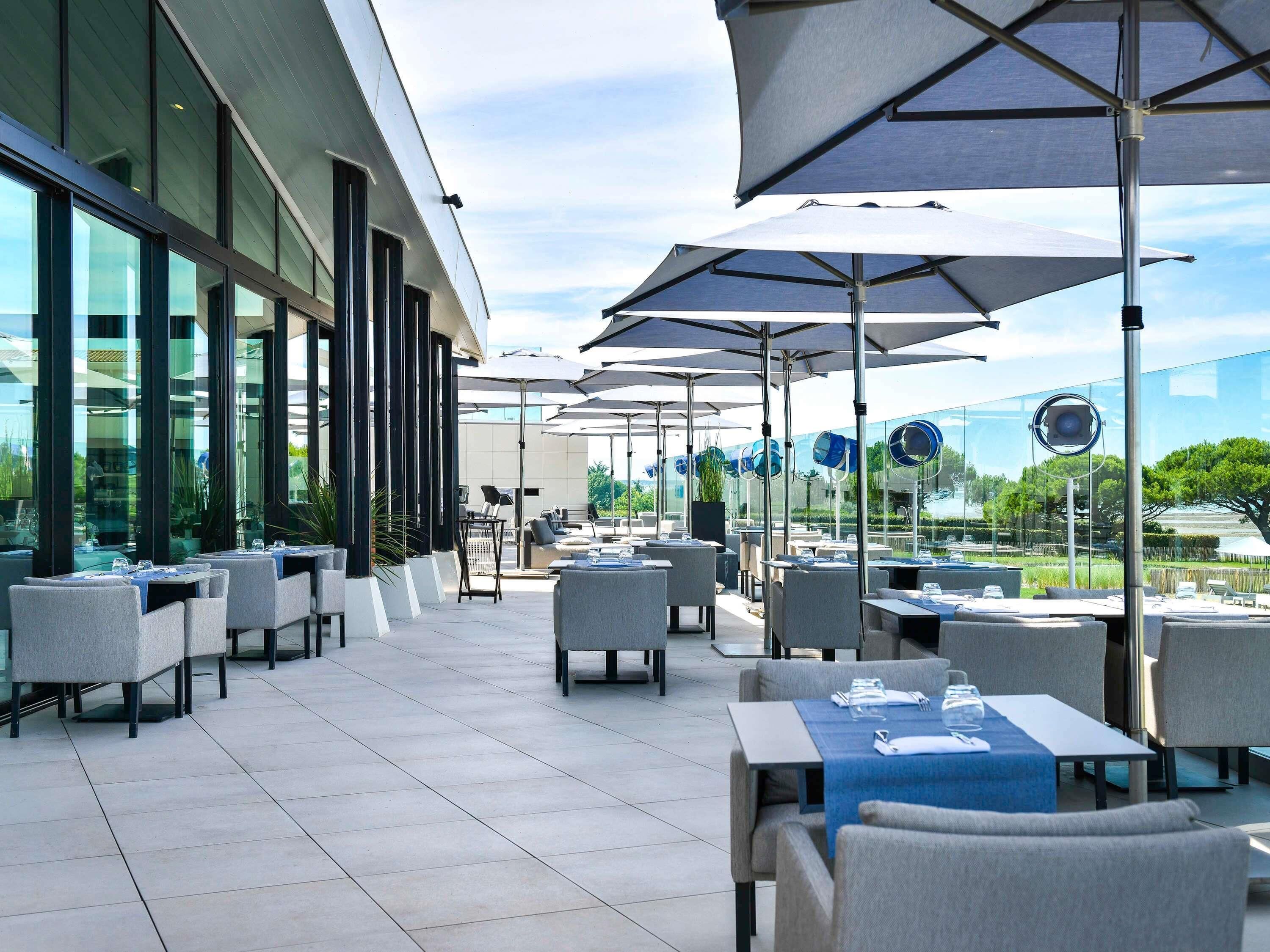 An outdoor dining area with tables and chairs, large umbrellas, and a glass building in the background.