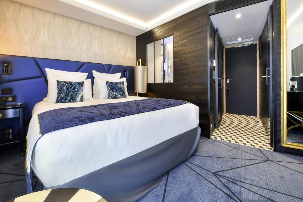 A modern hotel room with a large bed, a nightstand, a mirror, and a door leading to a bathroom.