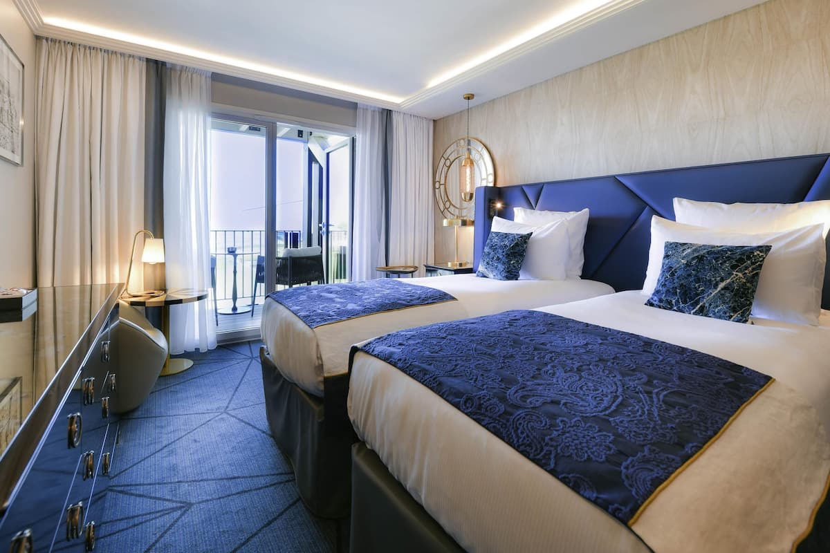 A hotel room with two beds, a desk, a chair, a lamp, and a view of the outside.