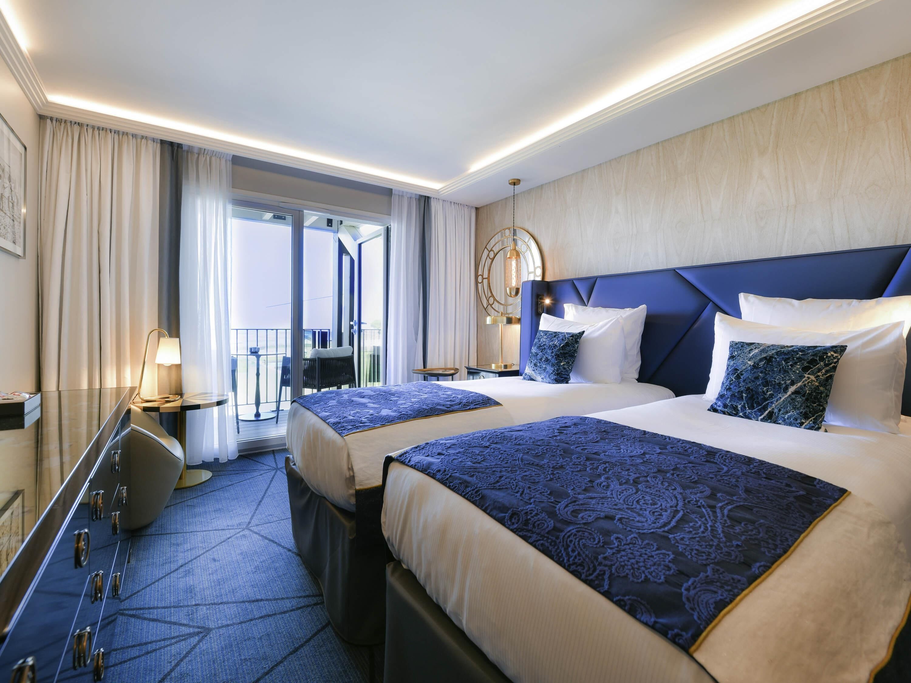 A hotel room with two beds, a desk, a chair, a lamp, and a view of the outside.