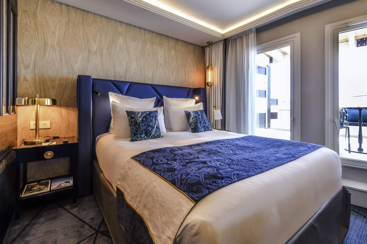 A hotel room with a large bed, a nightstand with a lamp, a wall-mounted TV, and a window with a view of the outside.