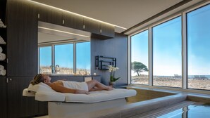 A spa room with a large window, a person relaxing on a massage table, and a view of the beach. - La Grande Terrasse Hotel & Spa La Rochelle - MGallery Collection (Chatelaillon-Plage)