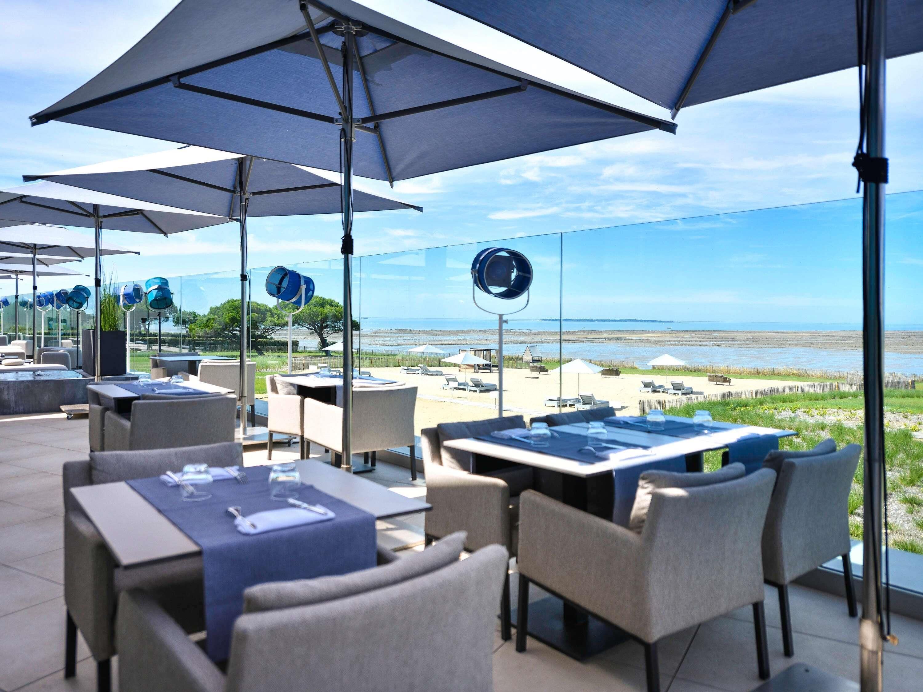 Un espacio al aire libre con mesas y sillas, grandes sombrillas y vistas a la playa.