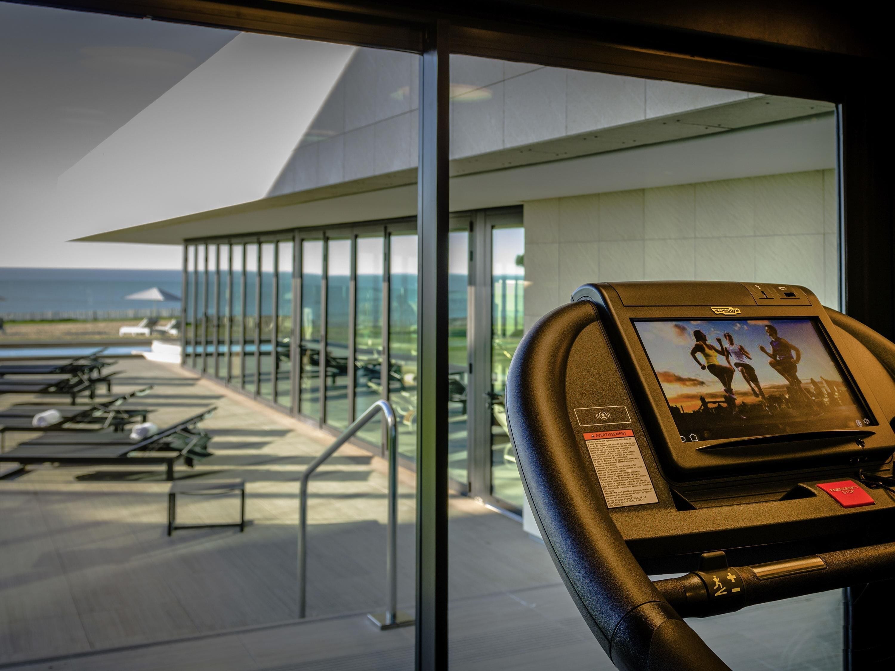 A treadmill with a digital display, overlooking a pool area with lounge chairs and a view of the ocean.