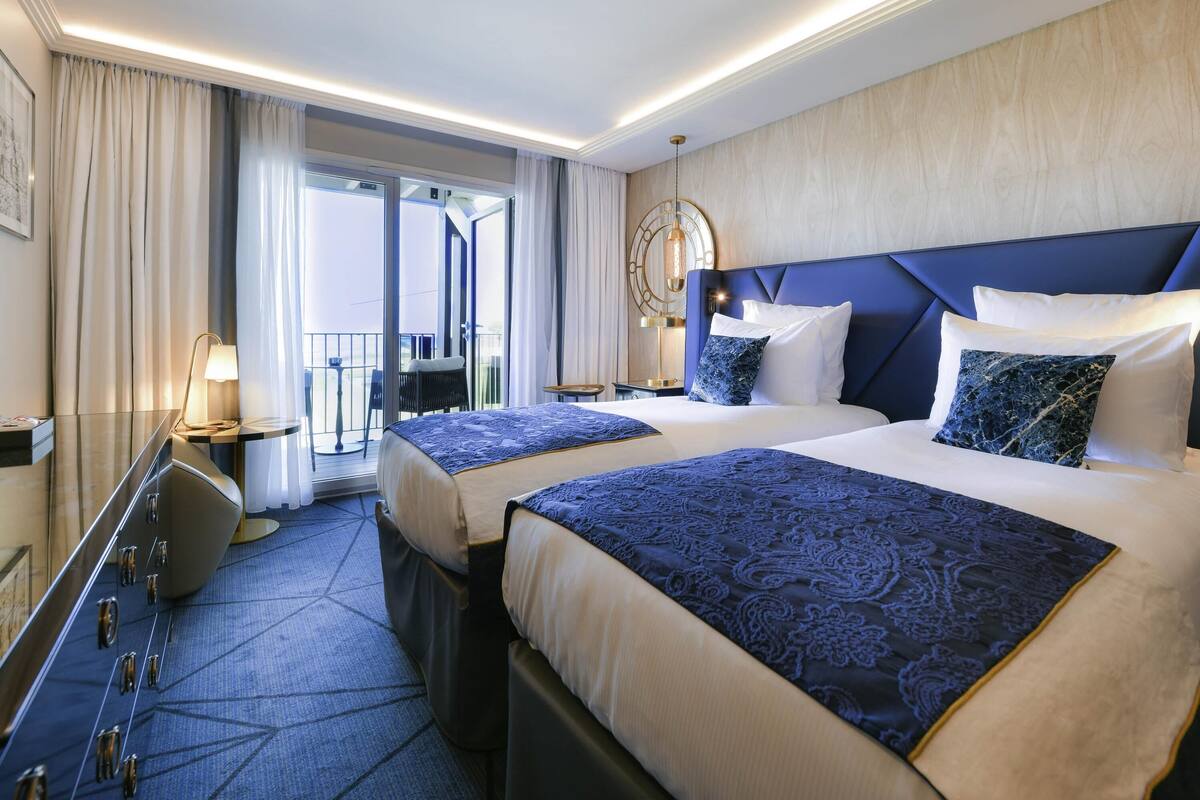A hotel room with two beds, a desk, a chair, a lamp, and a view of the outside.