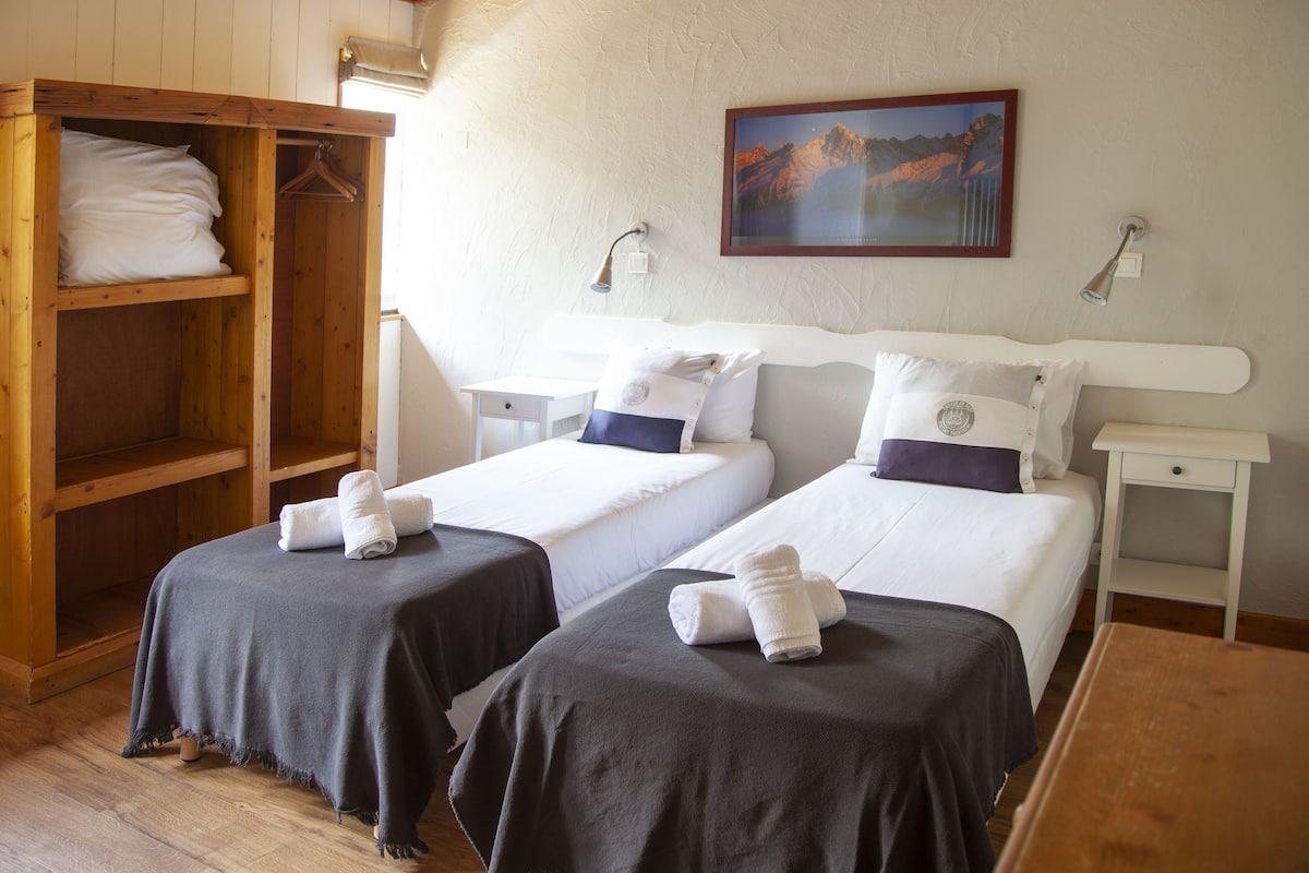 Habitación de hotel con dos camas, un armario de madera, un cuadro en la pared y una mesita de noche con lámpara.