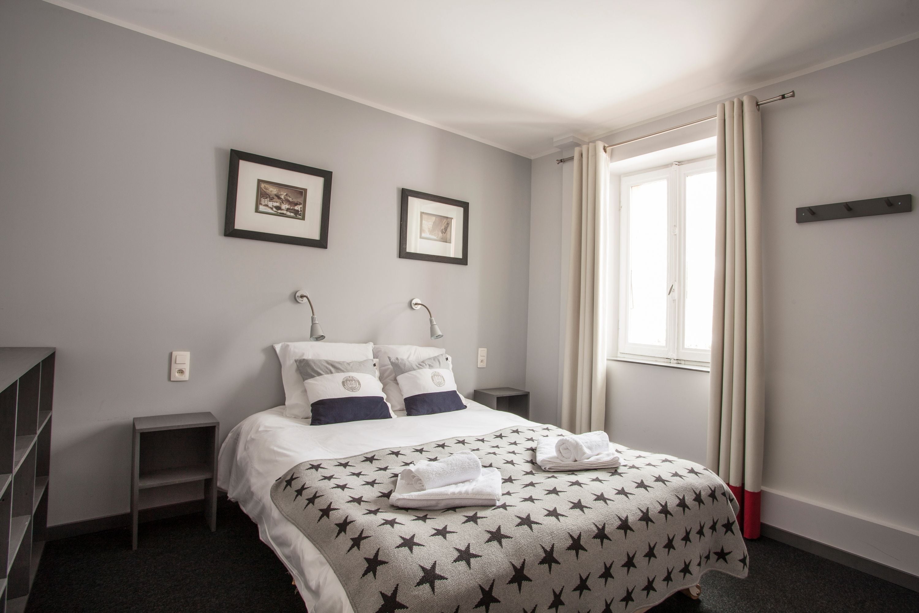 A neatly arranged hotel room with a bed, two bedside tables, a nightstand, a window with curtains, and two framed pictures on the wall.