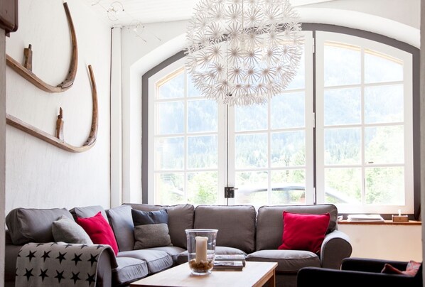 A modern living room with a sectional sofa, a glass table with a candle, and a large window with a view of a forest.