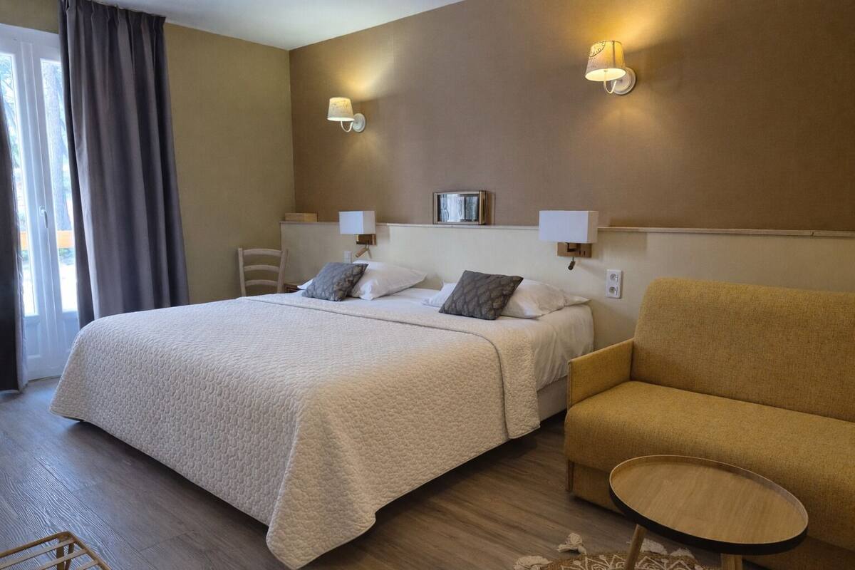 Ein Hotelzimmer mit einem Bett, zwei Nachttischlampen, einem Sessel, einem kleinen Tisch und Blick ins Freie.