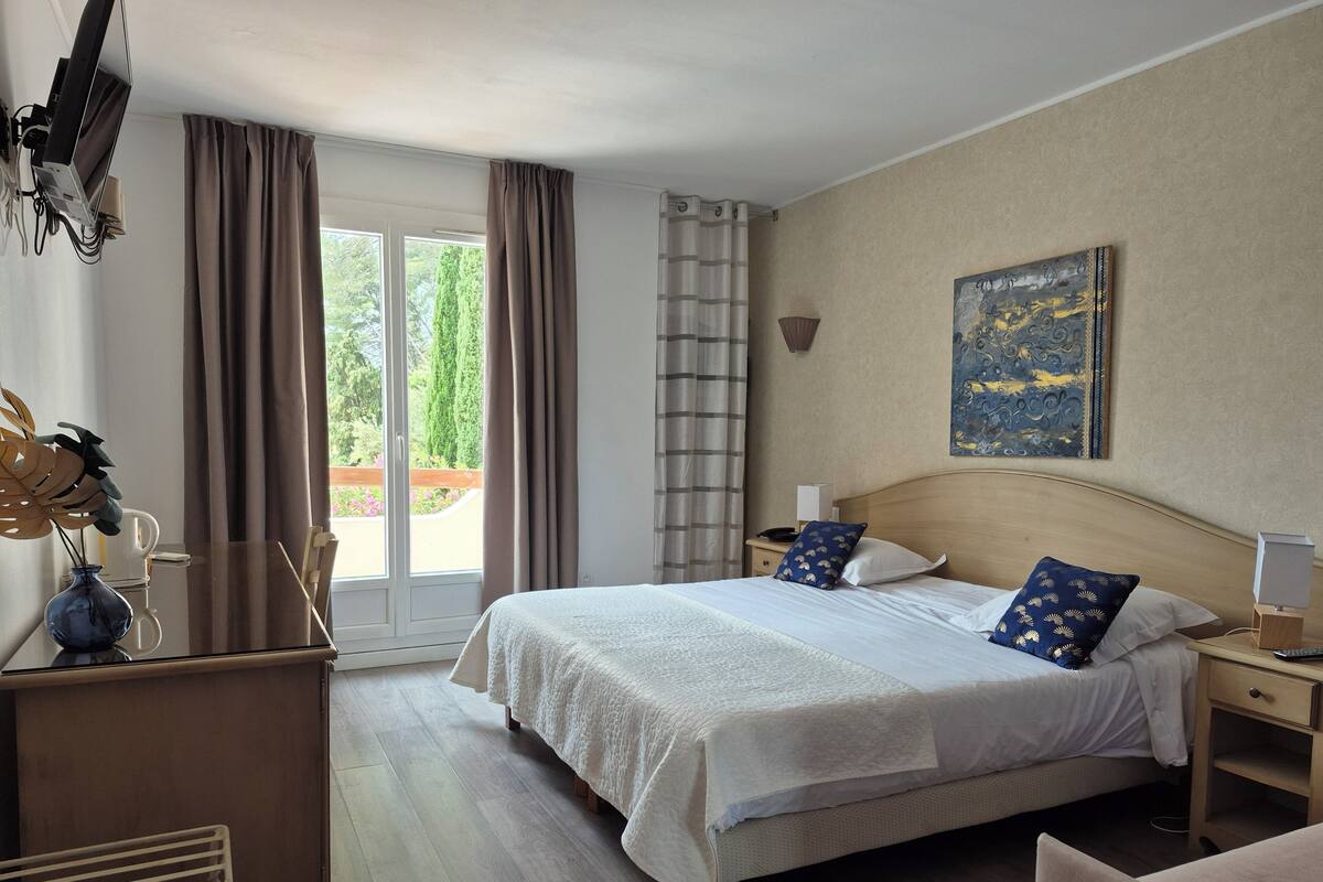 Ein Hotelzimmer mit einem Bett, Nachttischen, einem Schreibtisch mit Computer, einem Fenster mit Vorhängen und einem Bild an der Wand.