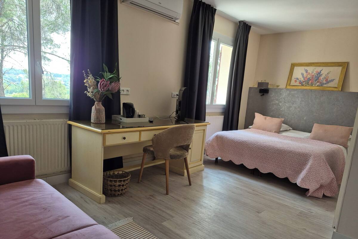 Ein Schlafzimmer mit Schreibtisch, einem Bett mit rosa Bettdecke, einem Sessel, einem Heizkörper und einem Fenster mit Vorhängen.