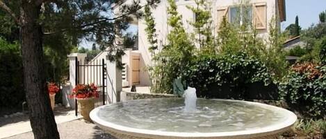 A circular water fountain with a stone base, surrounded by trees and a building in the background.