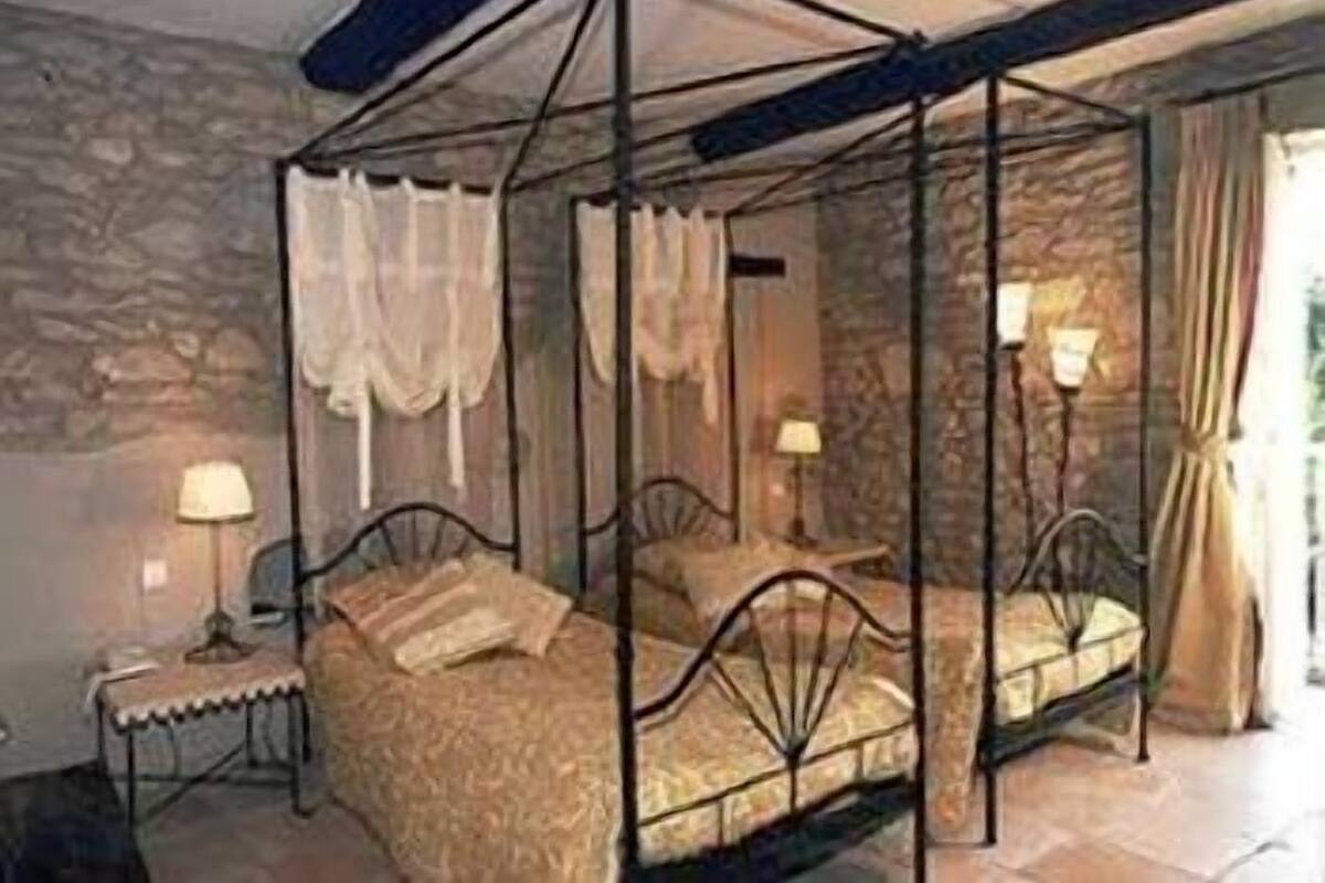 A four-poster canopy bed with a stone wall backdrop, two bedside tables with lamps, and a window with curtains.