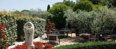 An outdoor seating area with tables and chairs, surrounded by well-maintained hedges and flowering plants.