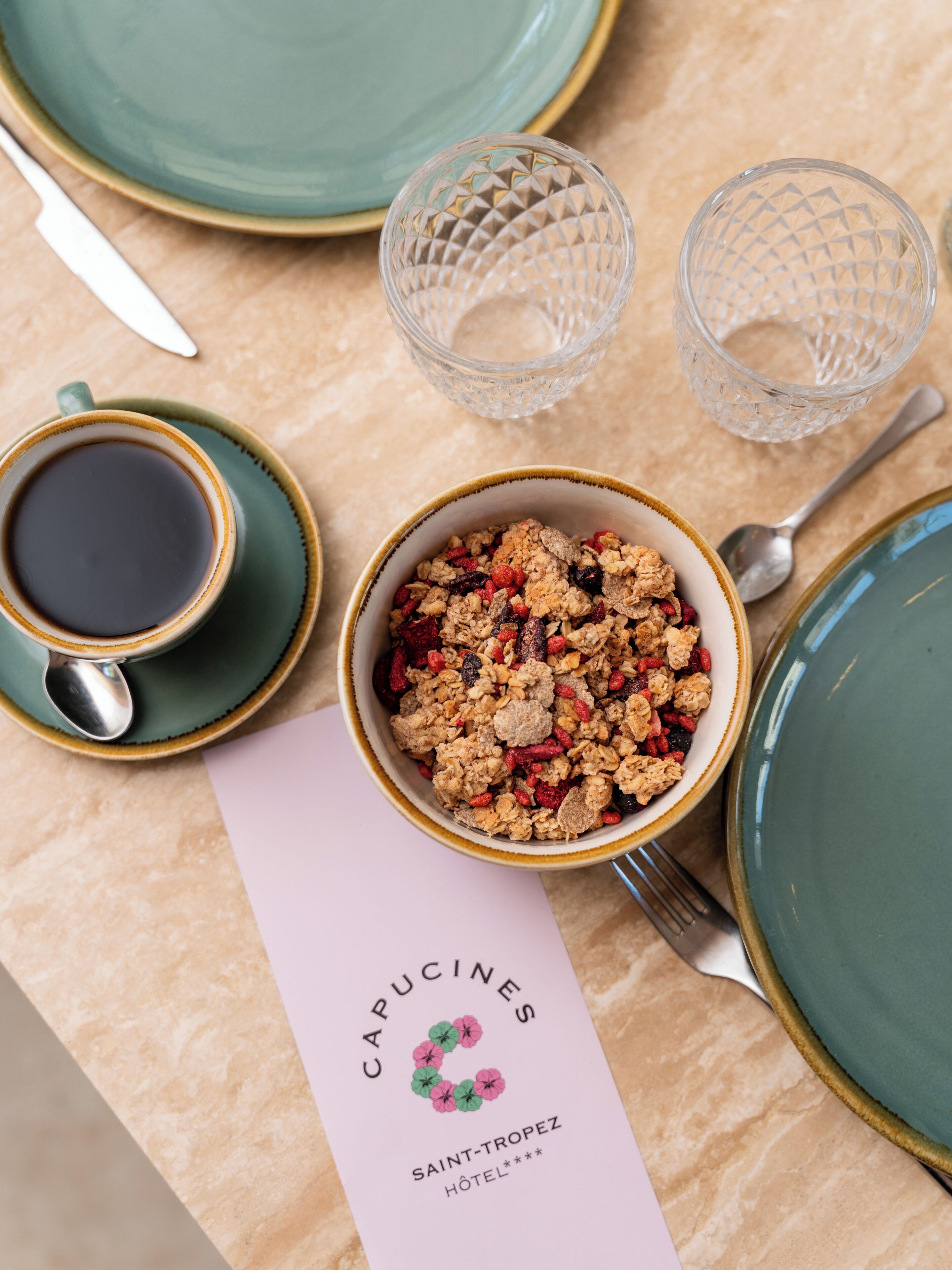 Une table dressée pour le petit-déjeuner avec un bol de granola, une tasse de café et un menu de l’hôtel Capucines Saint-Tropez.