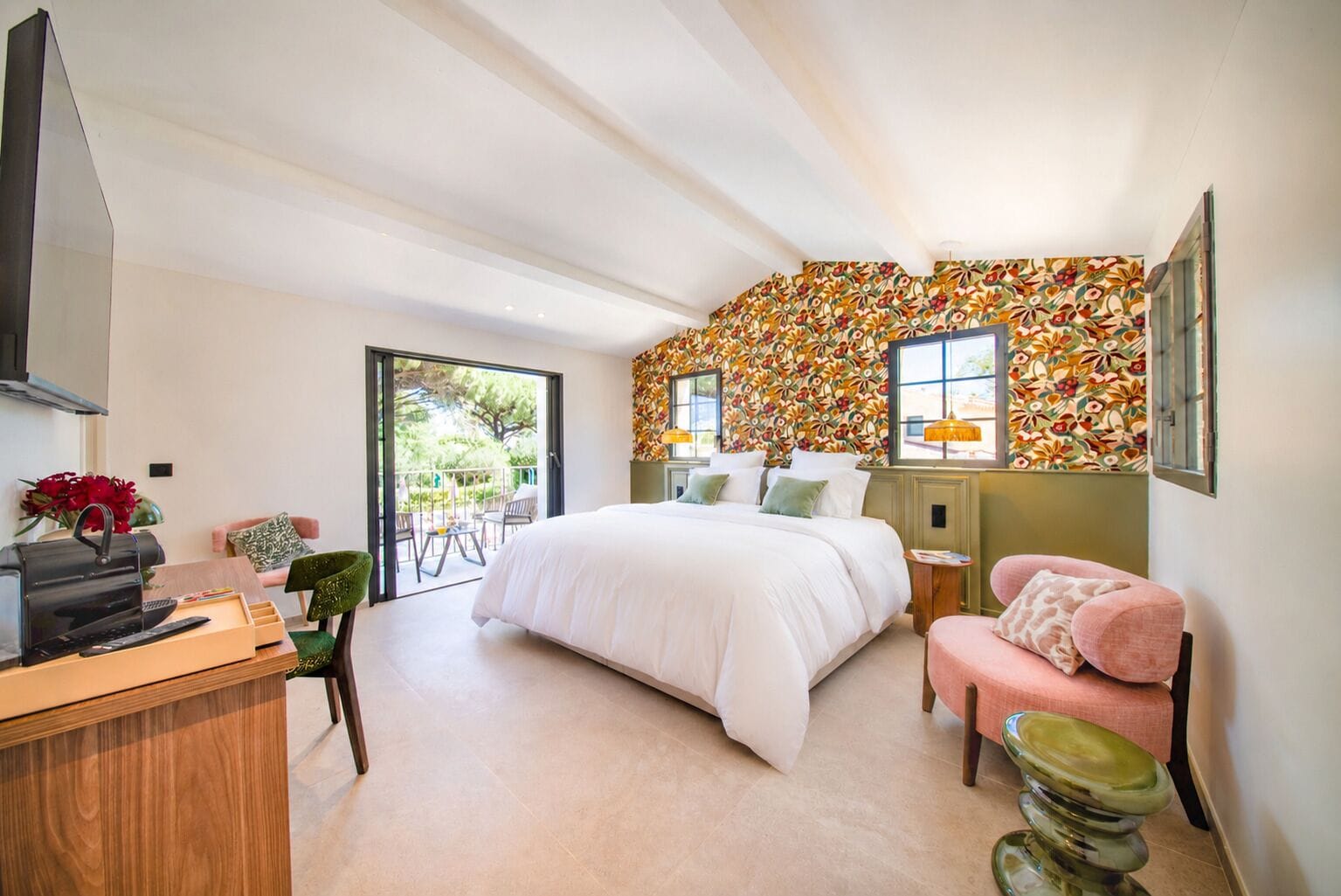 Ein Schlafzimmer mit einem großen Bett, einem Schreibtisch mit Computer, einem Sessel, einem rosa Sofa und einem Balkon mit Blick auf Grünflächen.