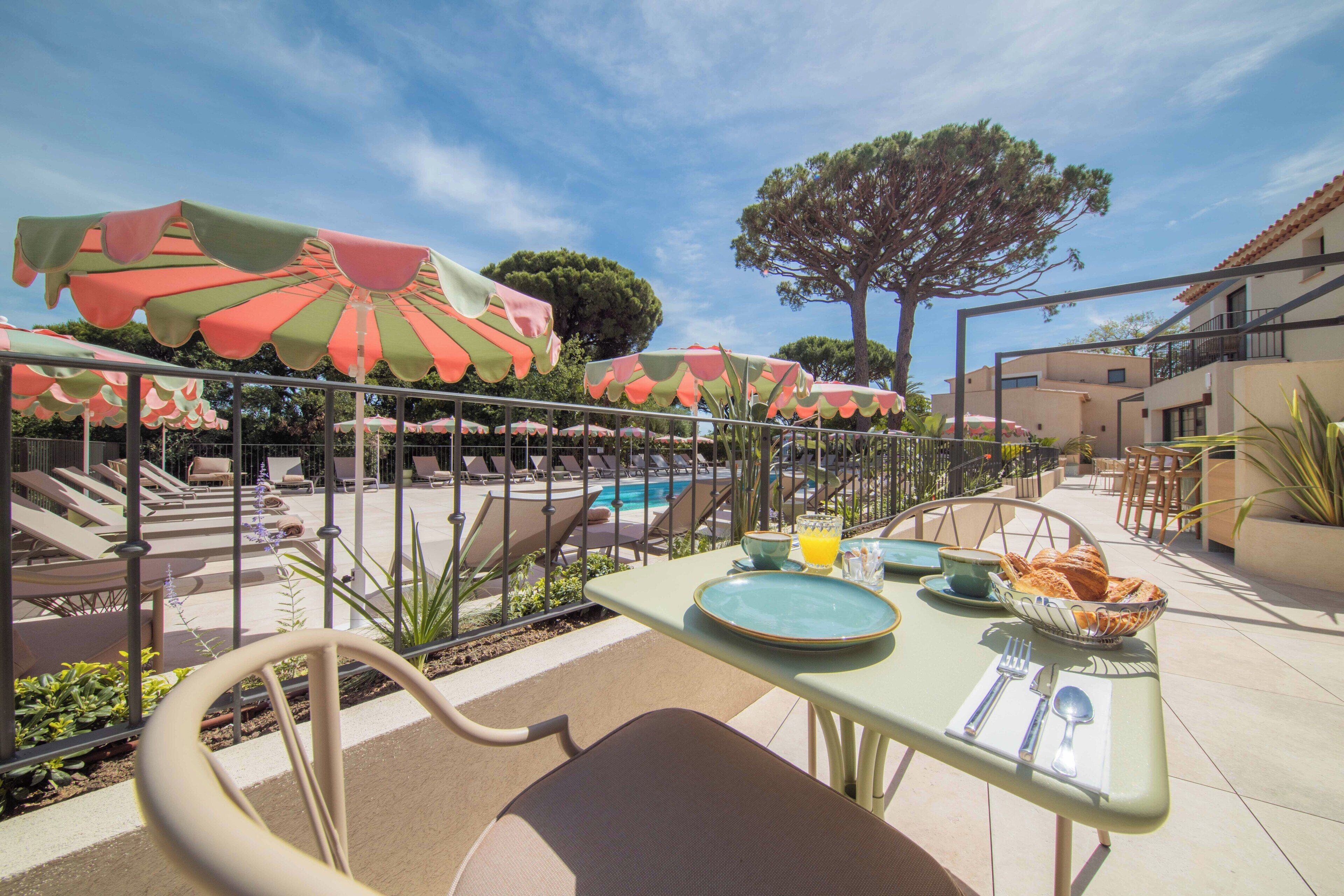 An outdoor dining area with tables, chairs, umbrellas, and a pool in the background.