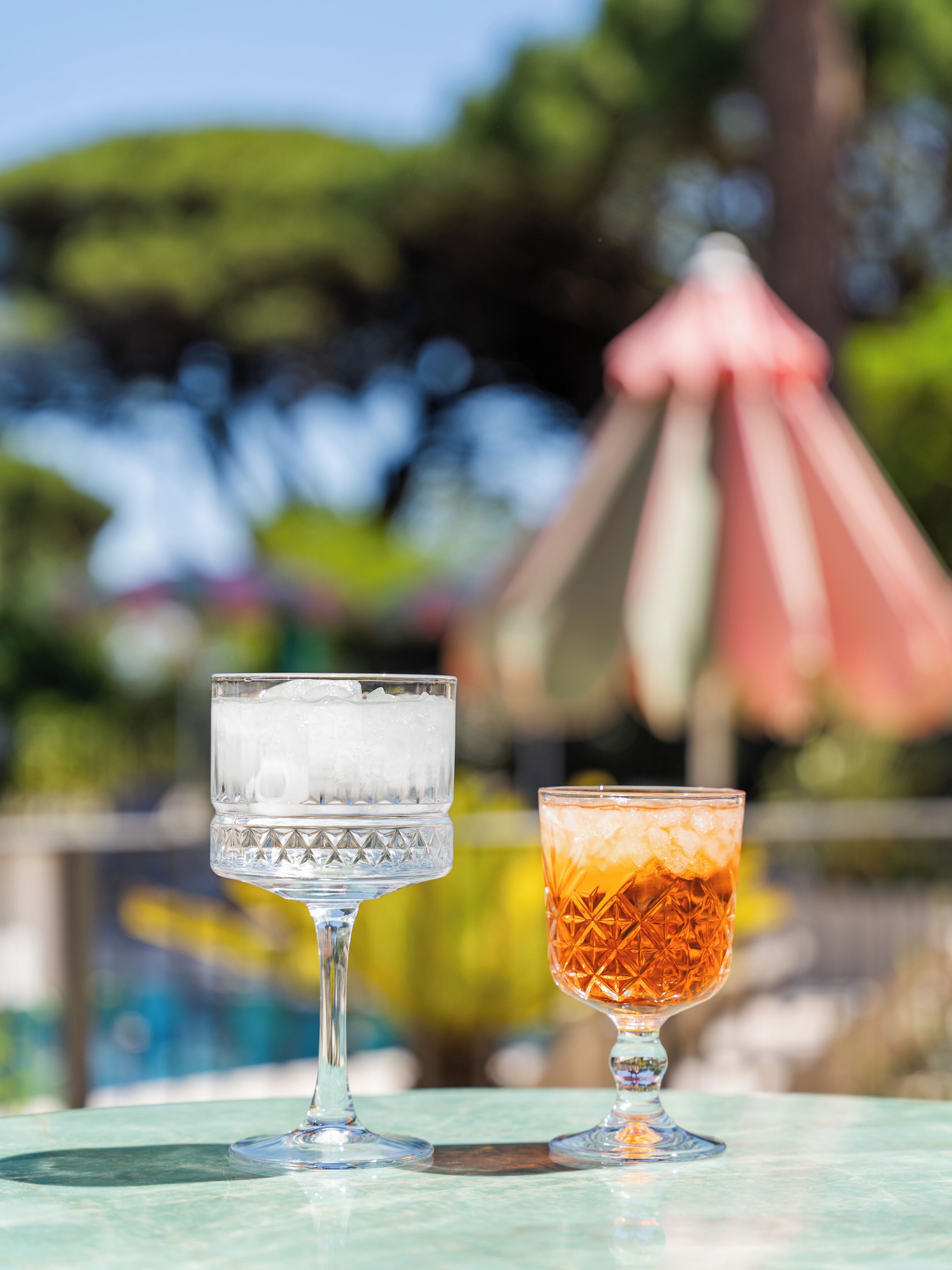 Deux verres de boissons glacées sont posés sur une table, avec une piscine et un parasol rose en arrière-plan.