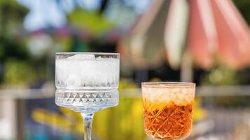 Two glasses of iced beverages on a table with a pool and a pink umbrella in the background.