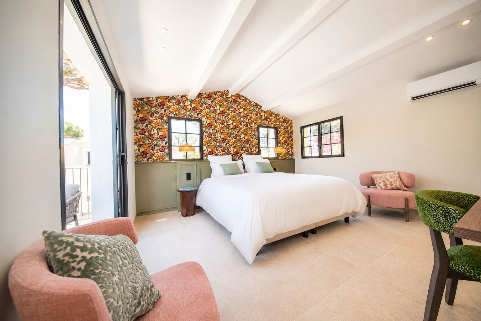 A bedroom with a large bed, two bedside tables, a chair, and a sofa. There are windows and an air conditioning unit.