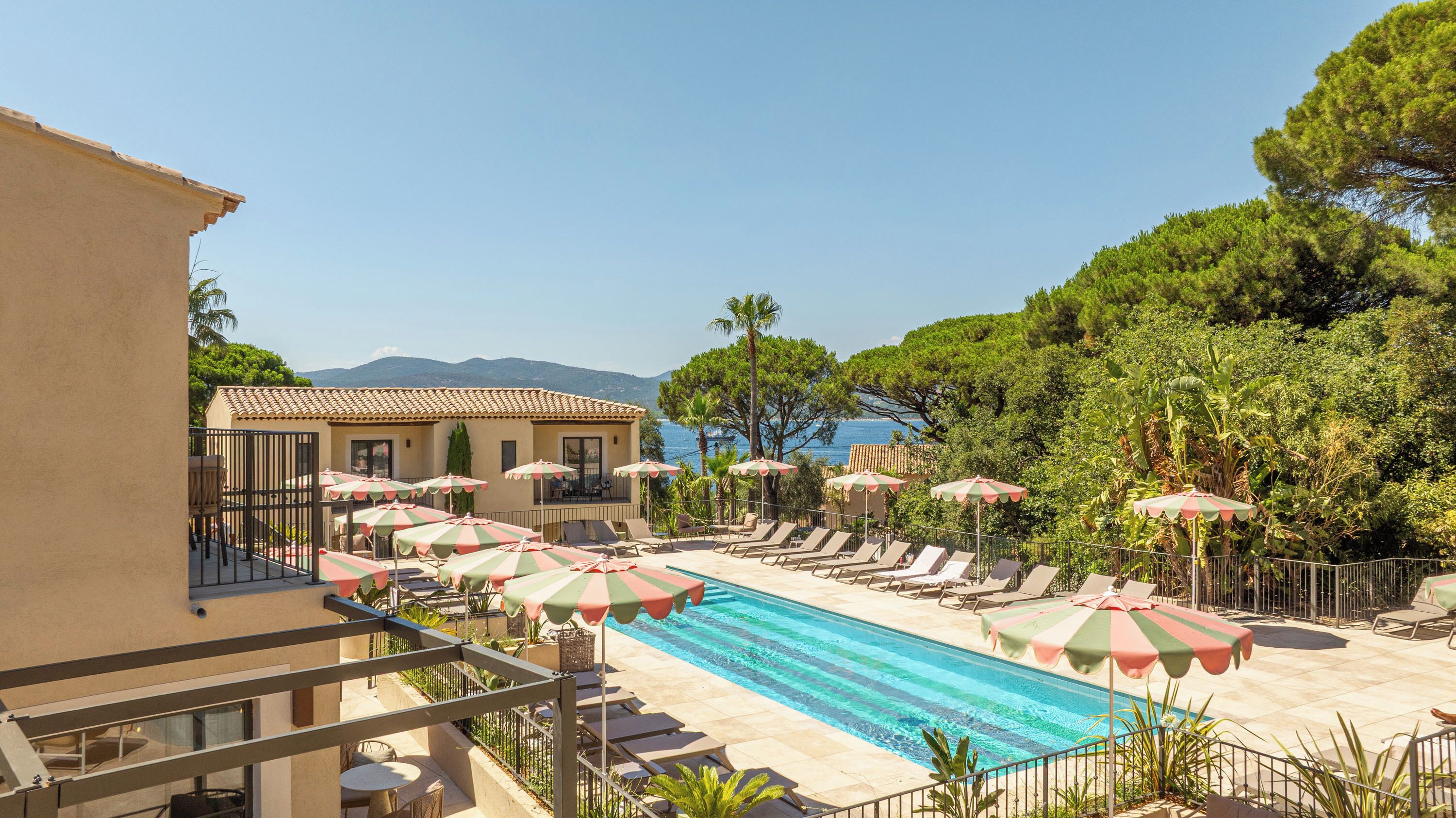 Un espace piscine avec des chaises longues et des parasols, un bâtiment doté d’un toit en tuiles, et une vue sur la mer et les montagnes.