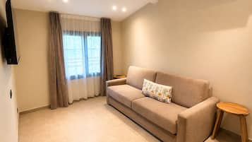 A living room with a sofa, a small side table, a TV mounted on the wall, and a window with curtains.