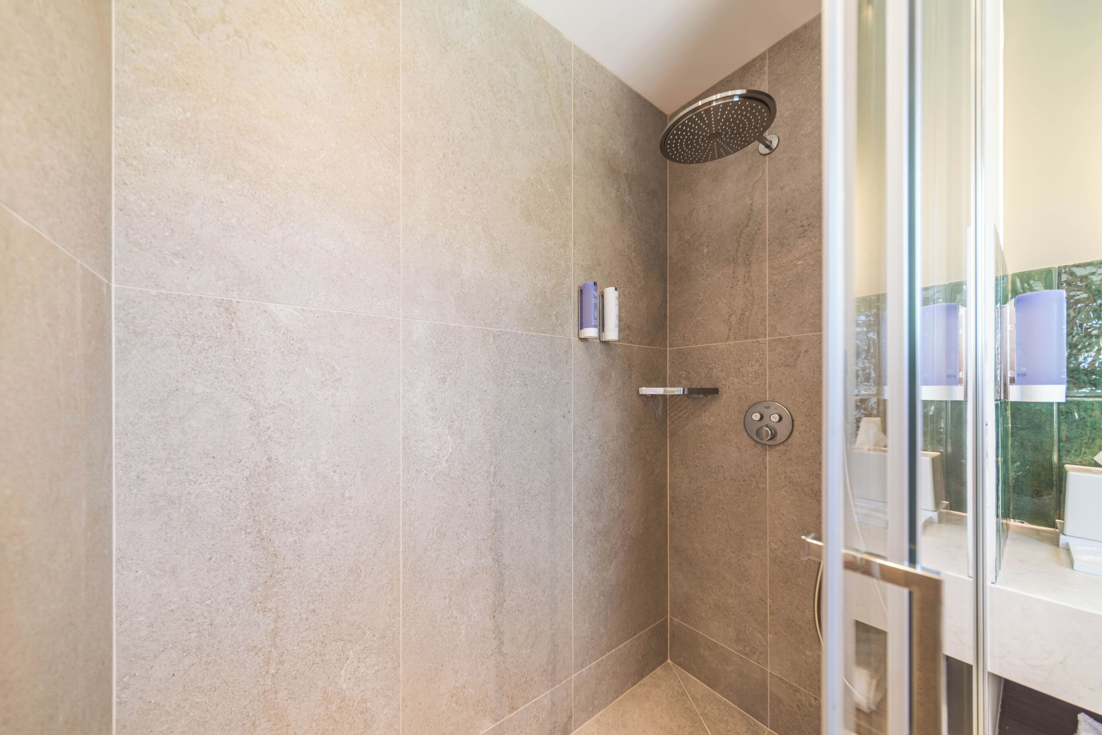Un baño moderno con baldosas color beige, mampara de vidrio y un dispensador de papel higiénico a la vista.