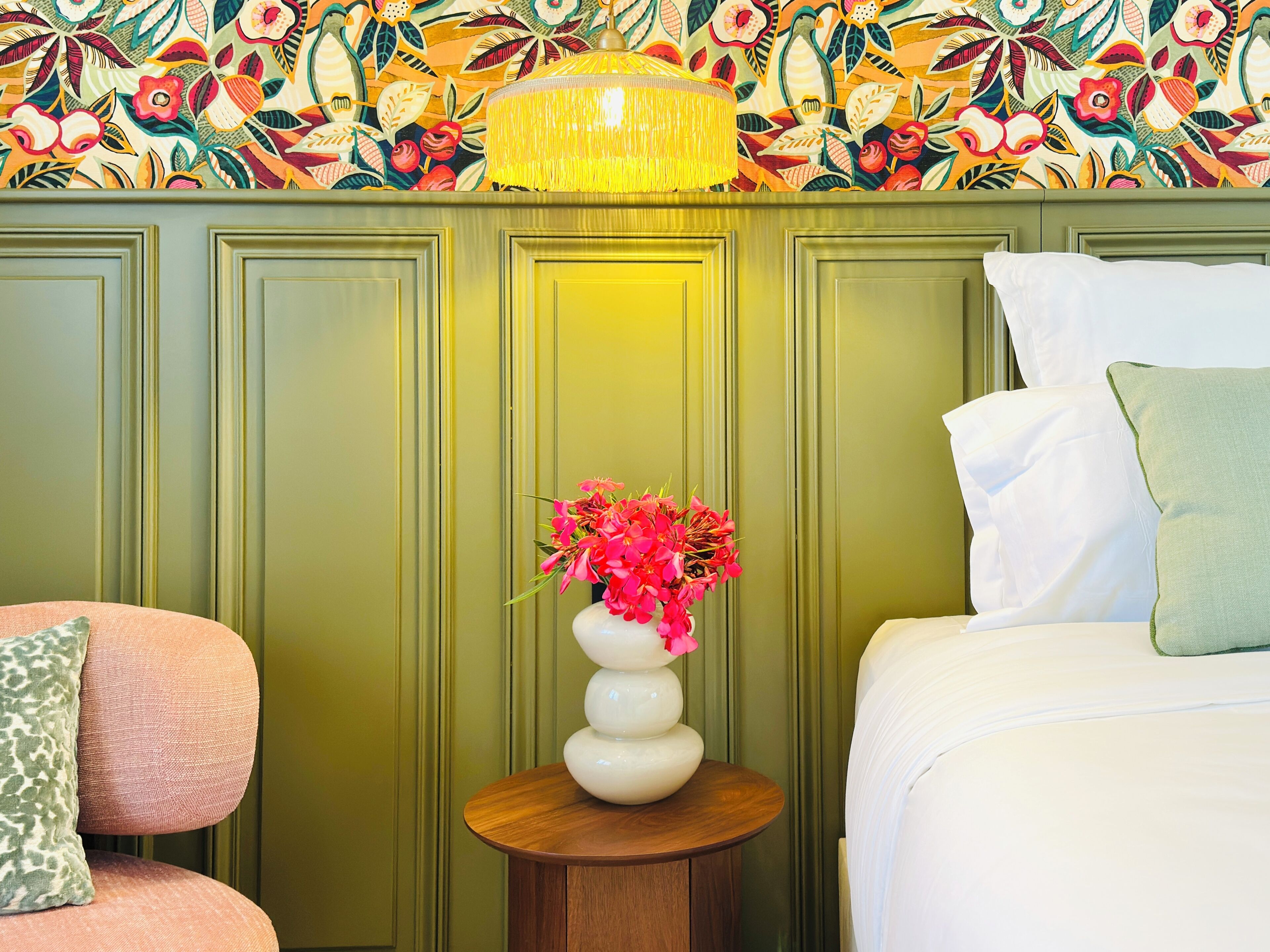 Ein Zimmer mit floralem Wandpapier, einem runden Holztisch mit Blumenvase, einem Bett mit weißer Bettwäsche und einem grünen Polsterstuhl.