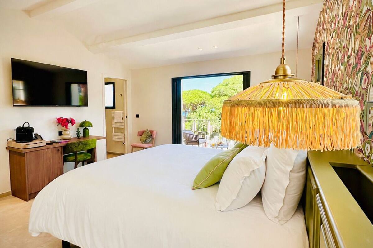 A bedroom with a large bed, a desk, a television, and a view of greenery outside.