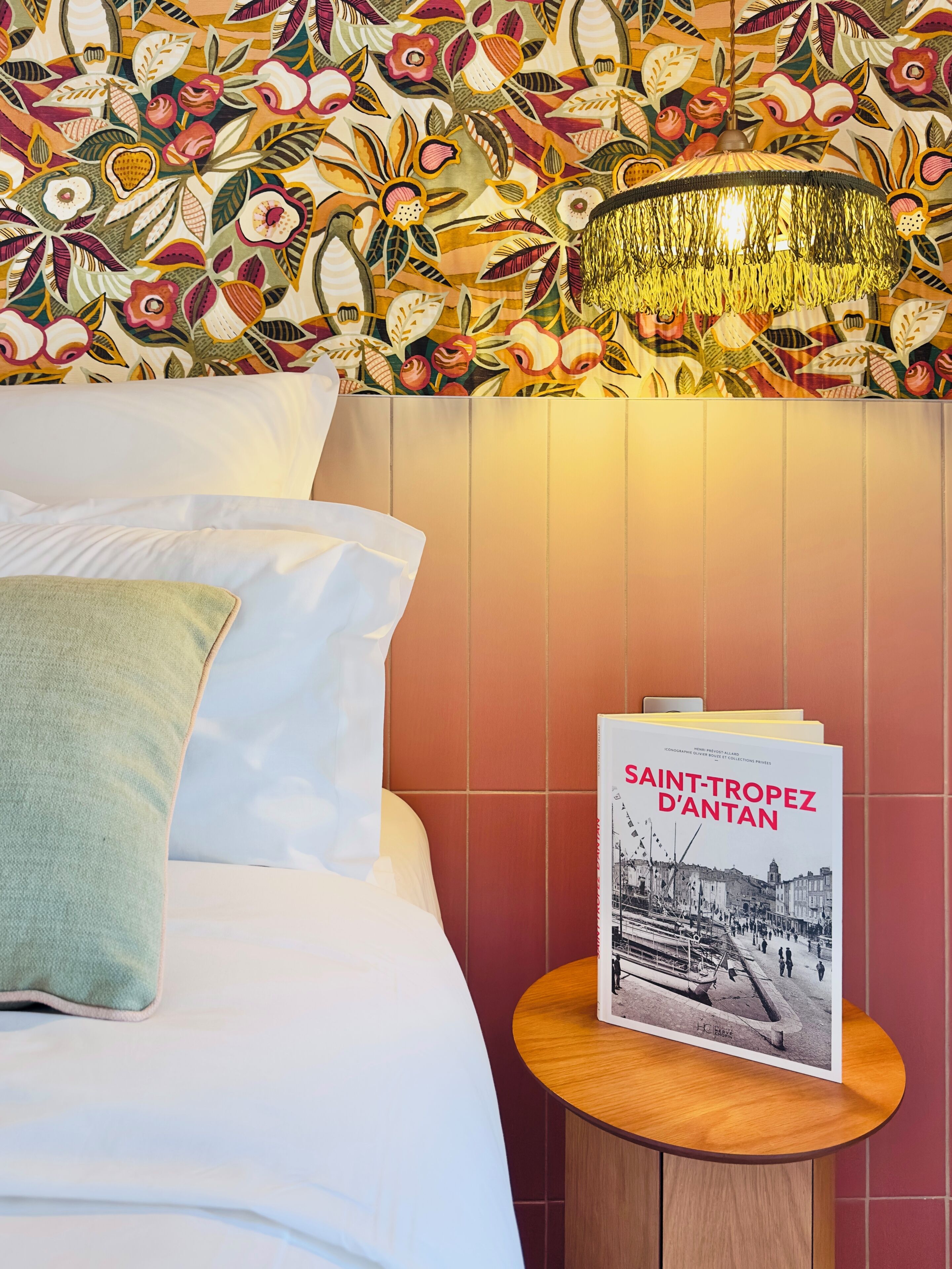 A hotel room with a bed, pillows, a round wooden nightstand, a book titled 'SAINT-TROPEZ D'ANTAN,' and a wall with a floral pattern.