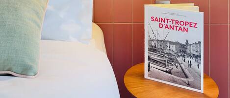 Ein Hotelzimmer mit einem Bett, Kissen, einem runden Nachttisch aus Holz, einem Buch mit dem Titel 'SAINT-TROPEZ D'ANTAN' und einer Wand mit floralem Muster.