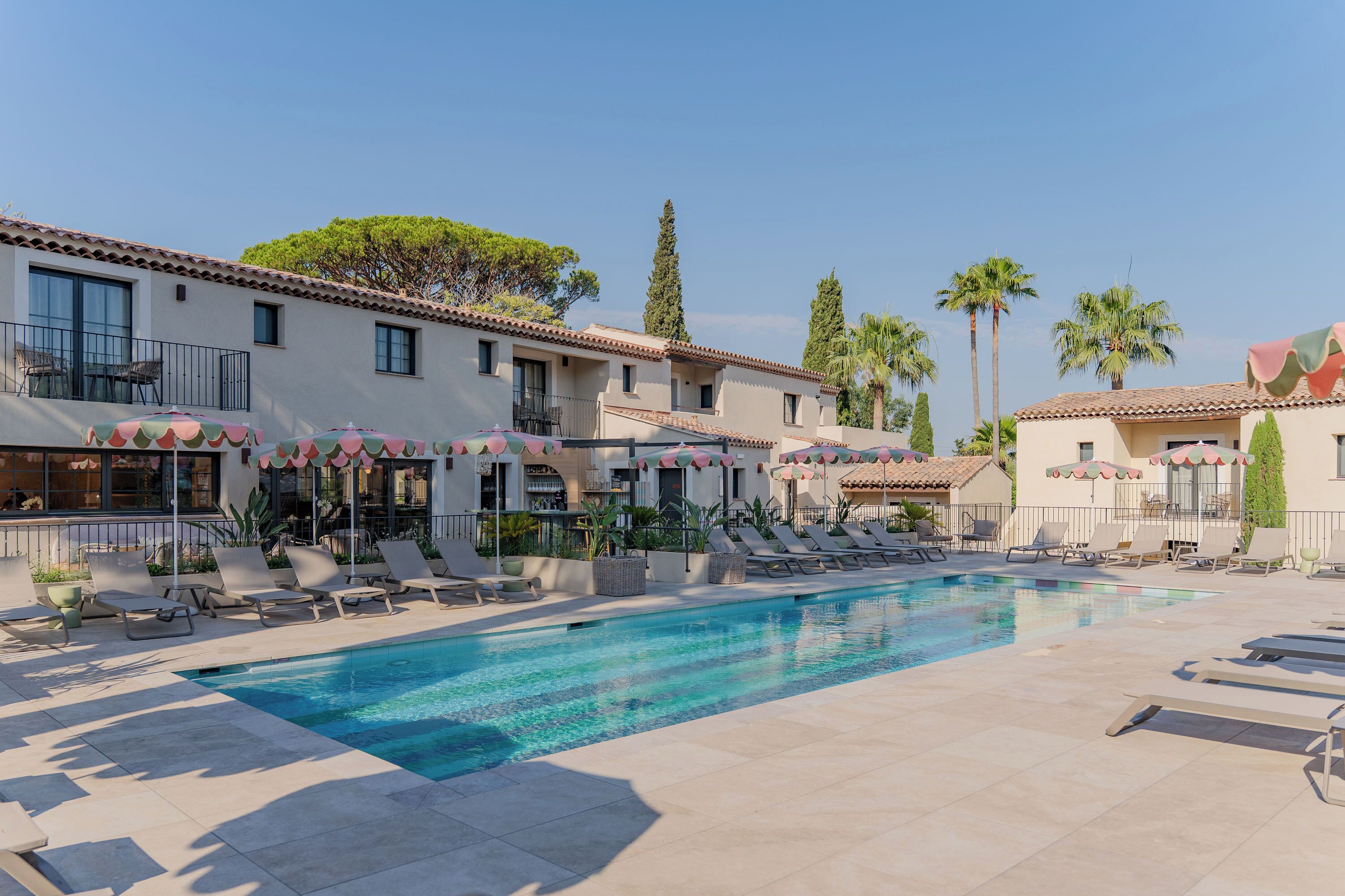Un espace piscine doté de chaises longues et de parasols, une piscine à l’eau d’un bleu limpide, et un bâtiment avec de nombreuses fenêtres et des balcons.
