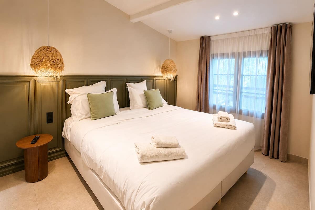 A hotel room with a large bed, two bedside lamps, a bedside table with a remote control, and a window with curtains.