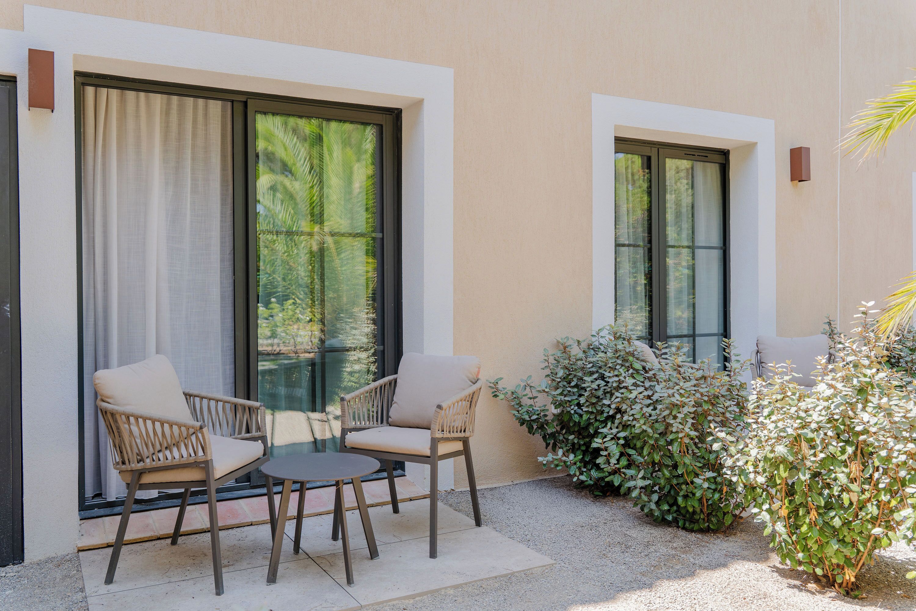 Un espace extérieur aménagé avec deux fauteuils en osier et une petite table, un pot de fleurs et une vue sur la verdure par une fenêtre.
