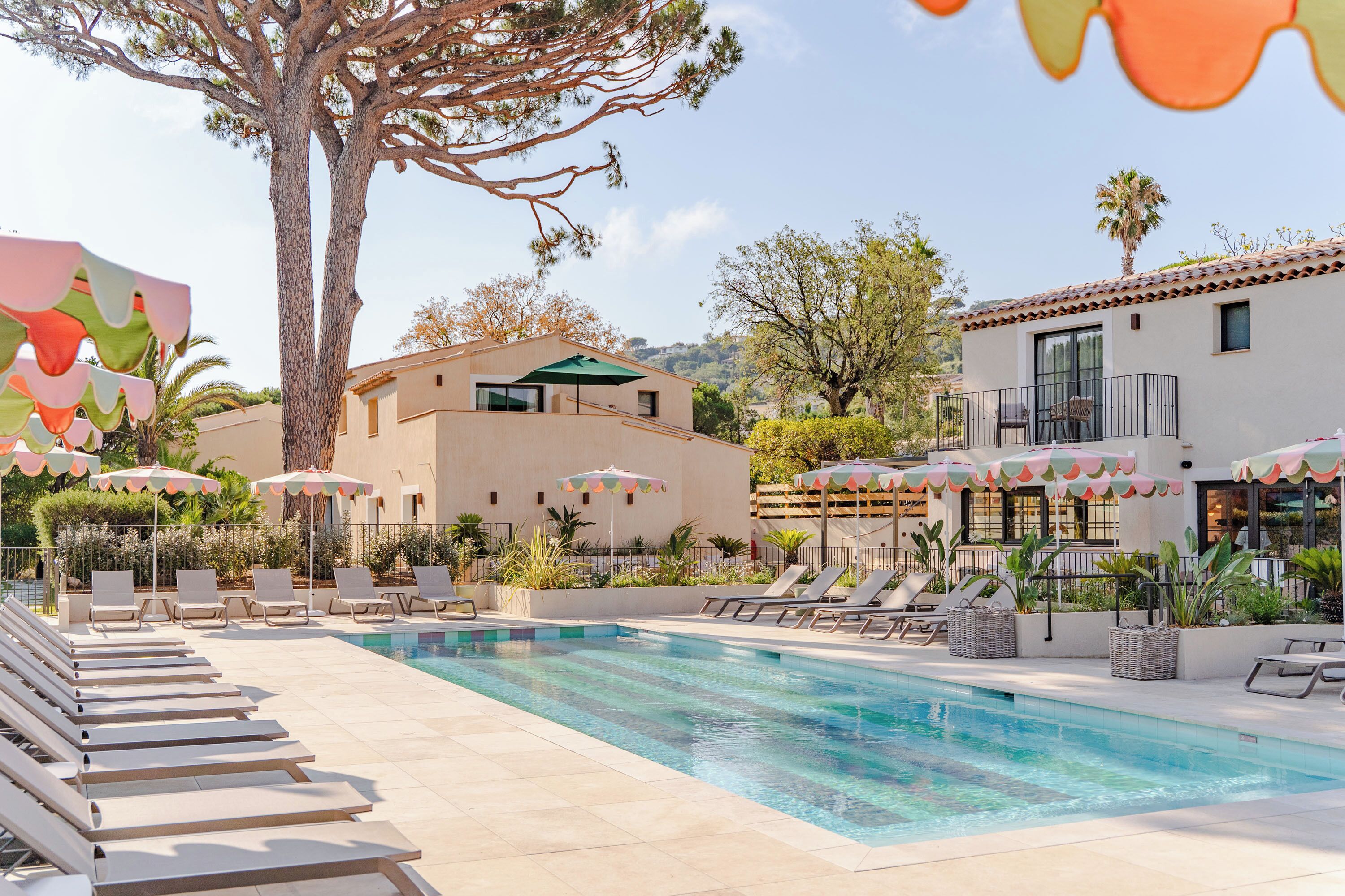 Un área de piscina con sillones de playa y sombrillas, un edificio de diseño moderno y un árbol grande.