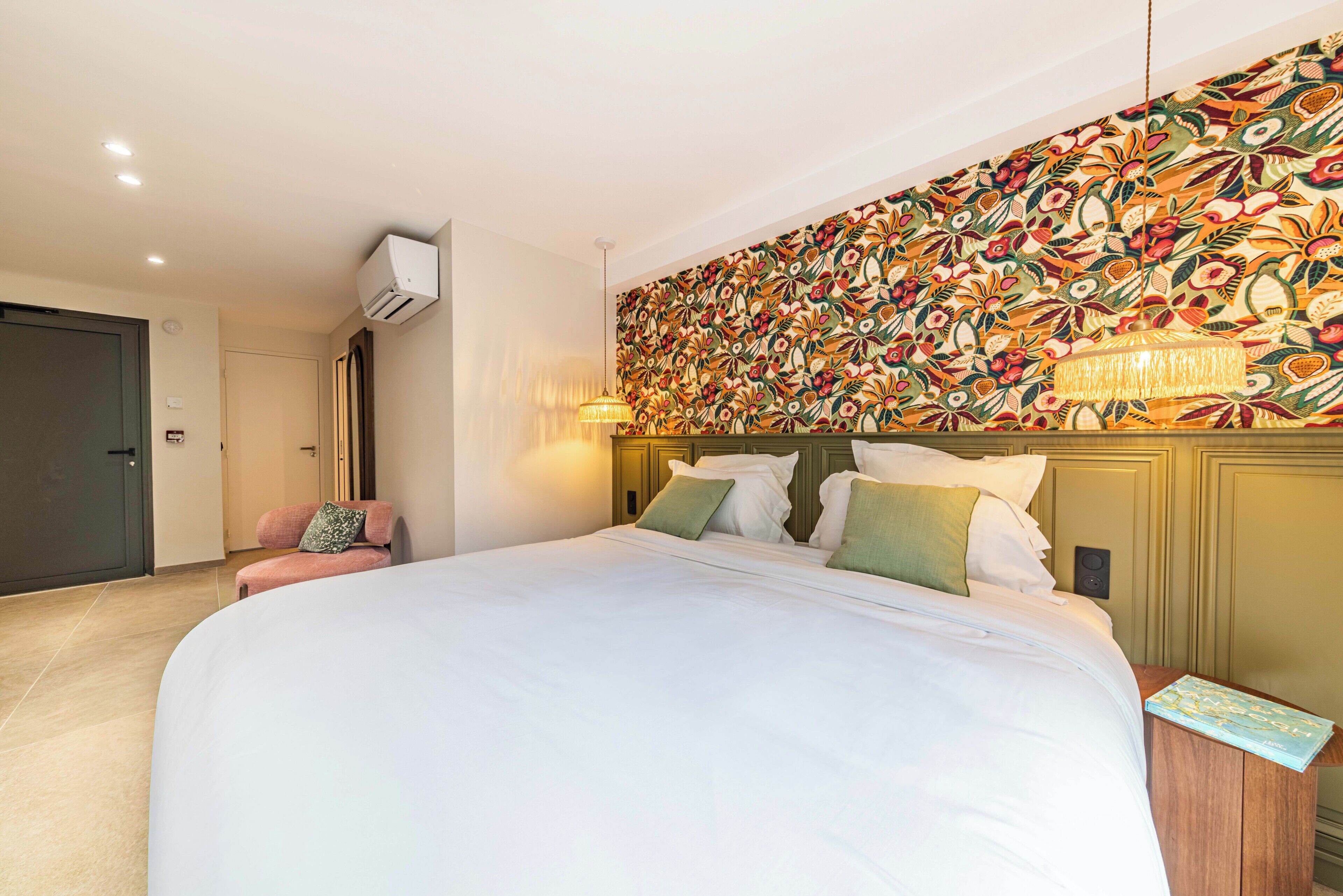 A hotel room with a large bed, a chair, a wall-mounted air conditioner, and a patterned wallpaper.