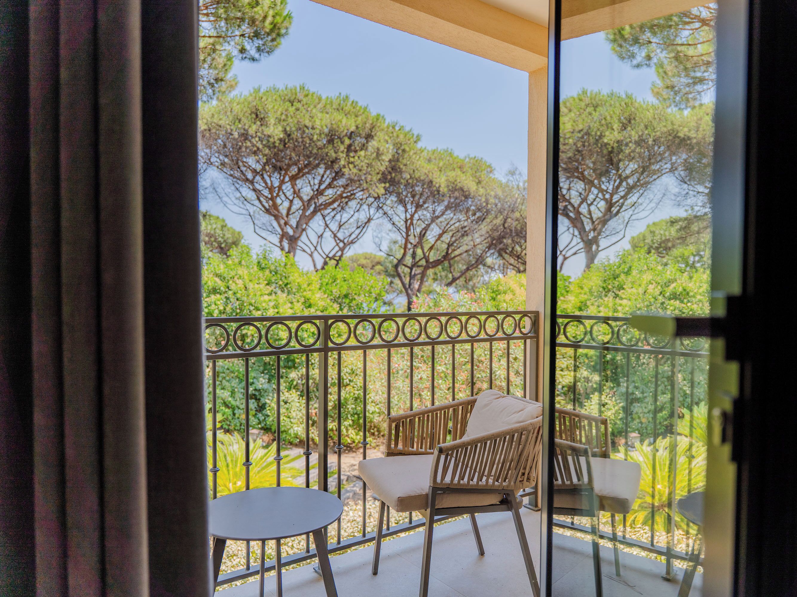 A balcony with two chairs and a small table, overlooking a garden with trees and shrubs.