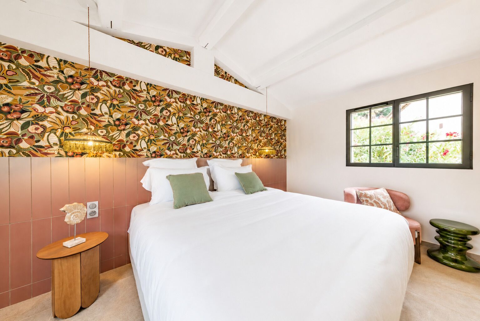 Un dormitorio con una cama grande, una lámpara de pared, una mesita de madera pequeña y una ventana con vistas a follaje.