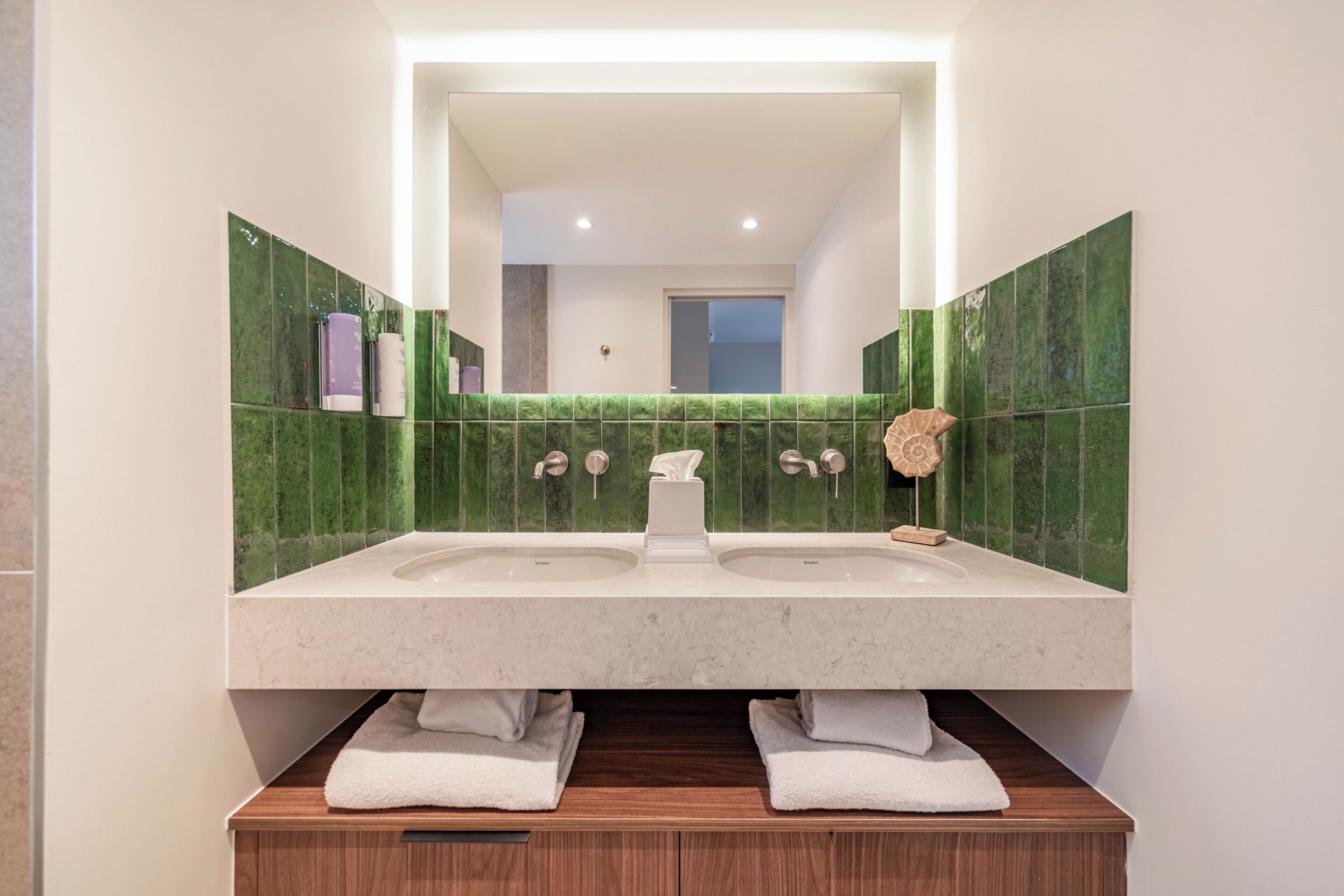 Un baño con lavamanos doble, azulejos verdes, un espejo y un mueble de tocador de madera.