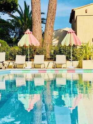 A swimming pool with lounge chairs and umbrellas, surrounded by palm trees and a building.