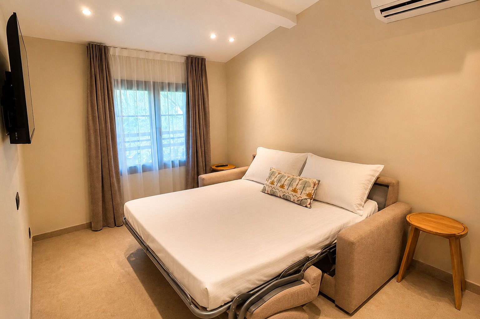 A hotel room with a bed, a TV, a bedside table, and a window with curtains.